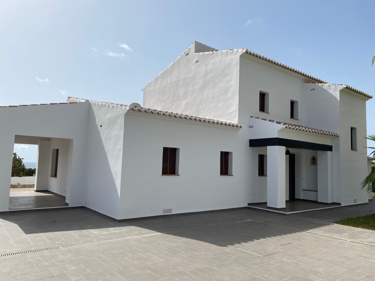 Fachada mediterránea con paredes blancas, tejados de teja curva y un amplio patio pavimentado en tonos grises con canales de drenaje integrados.