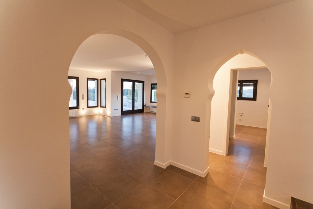 Pasillo con arcos decorativos de inspiración mediterránea, suelos de gres en tonos tierra y acceso al salón con ventanales de carpintería oscura.
