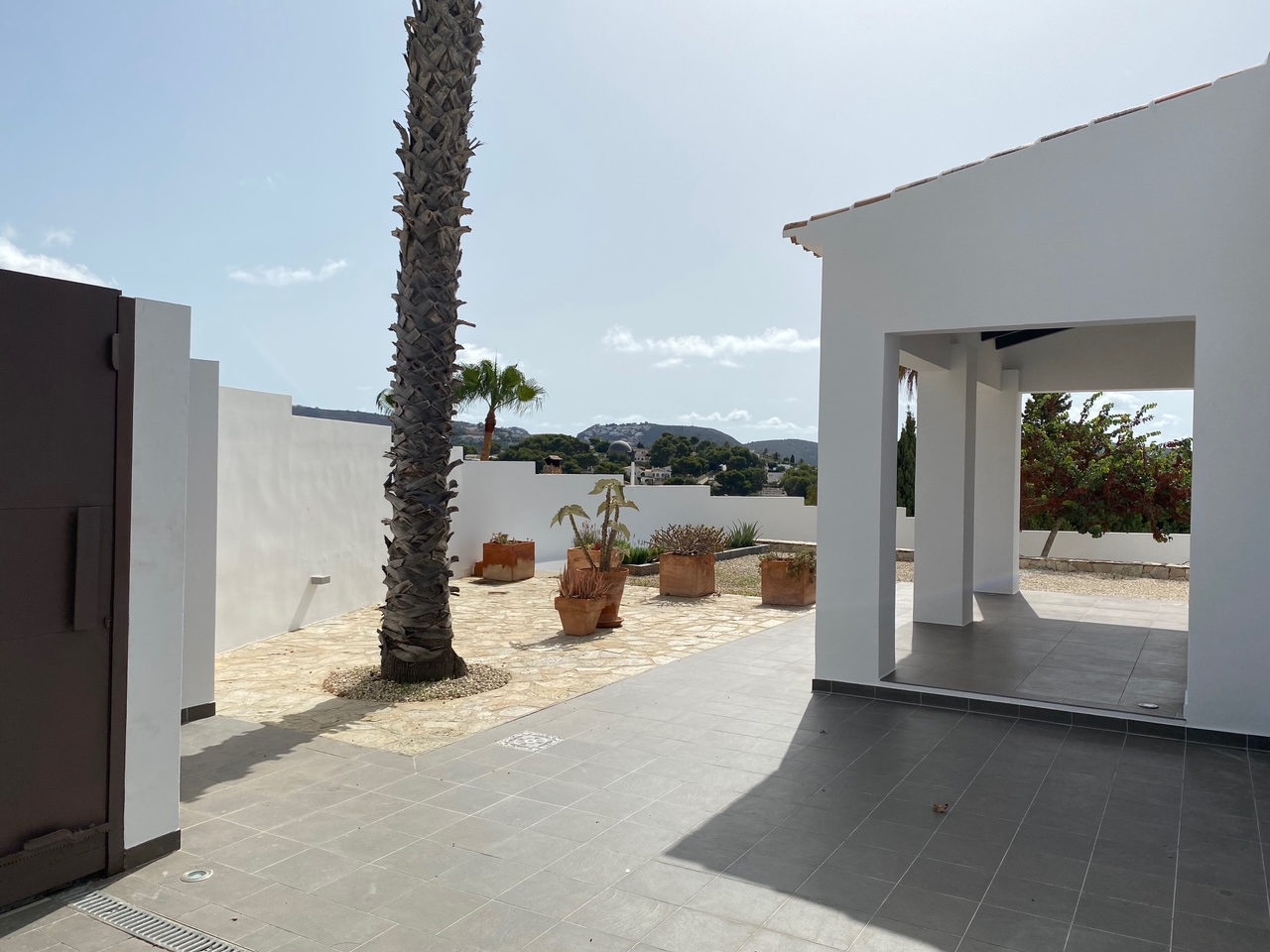 Patio exterior con suelos de gres gris y piedra natural, presidido por una palmera y macetas de terracota, con vistas despejadas a las colinas de Moraira.