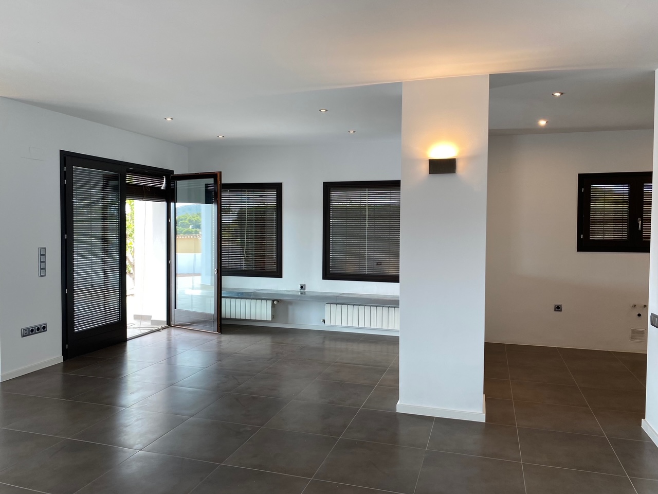 Salón diáfano con suelo de gres oscuro, iluminación empotrada y radiadores bajo ventanas con persianas venecianas. Incluye preinstalaciones técnicas y acceso directo al exterior.