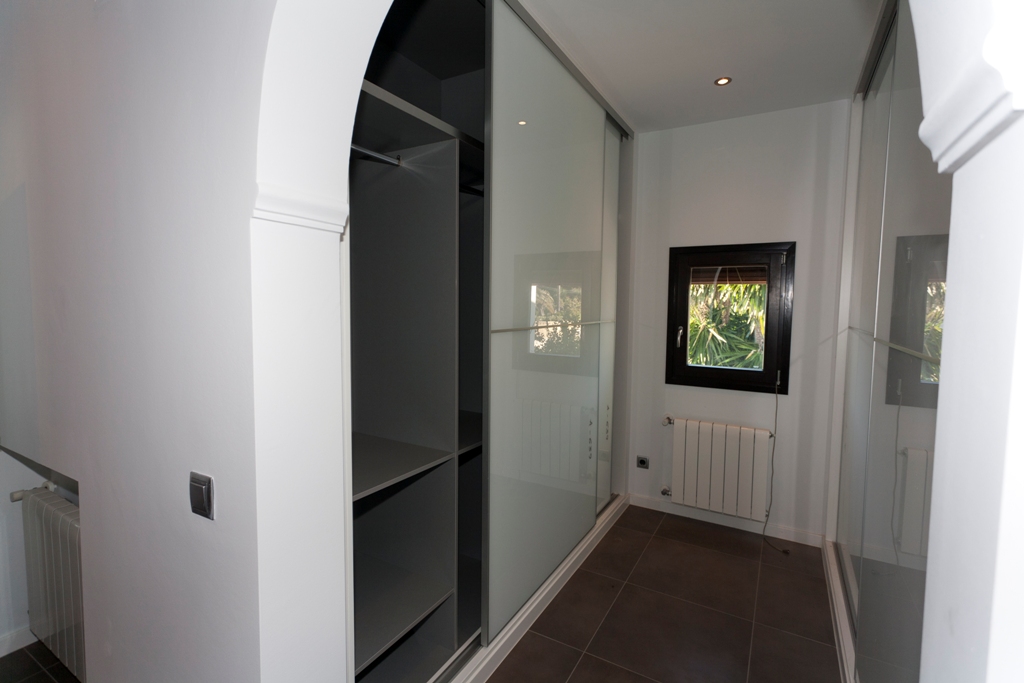 Vestidor con armarios empotrados de puertas correderas de cristal, estanterías interiores, suelos de gres oscuro, calefacción por radiador e iluminación empotrada.