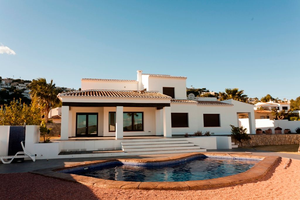 Vista exterior de la villa con piscina irregular, porche cubierto con vigas de madera oscura y fachada blanca de estilo mediterráneo tradicional.