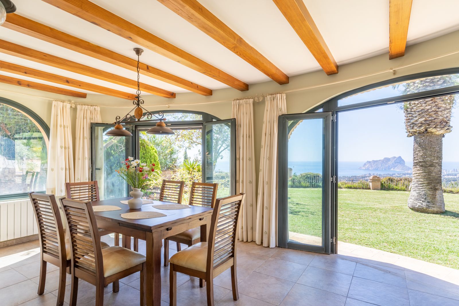 Comedor luminoso con vigas de madera y vistas al jardín y al mar Mediterráneo.