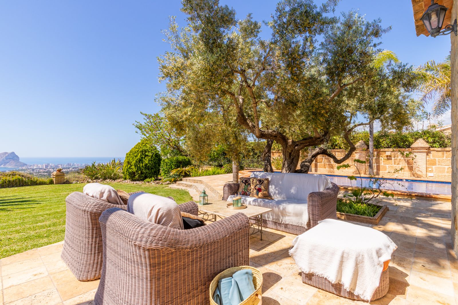 Terraza con muebles de mimbre y vistas al mar, bajo la sombra de un olivo centenario.
