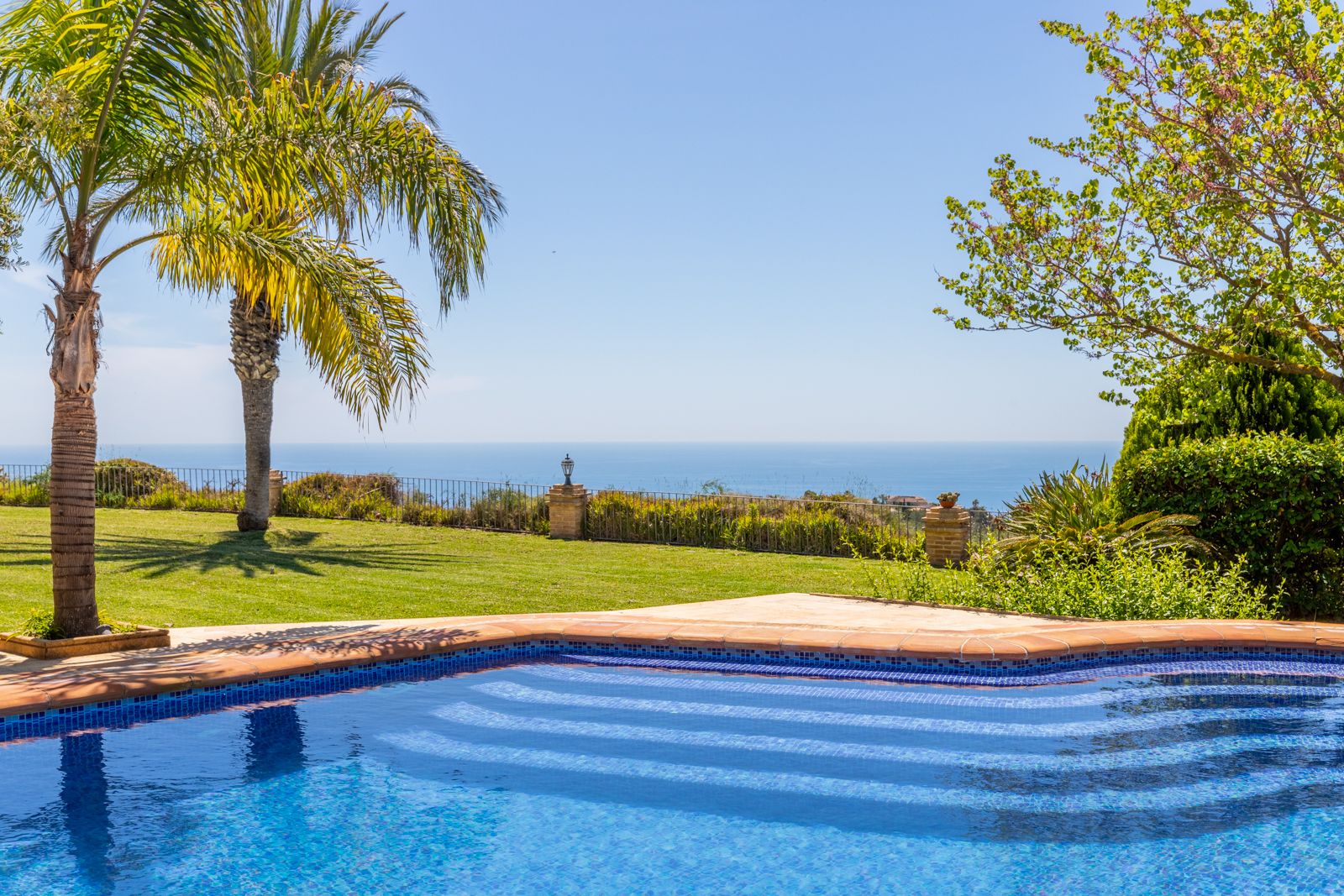 Vista de la piscina con mosaicos azules y el jardín con palmeras, frente al mar Mediterráneo.