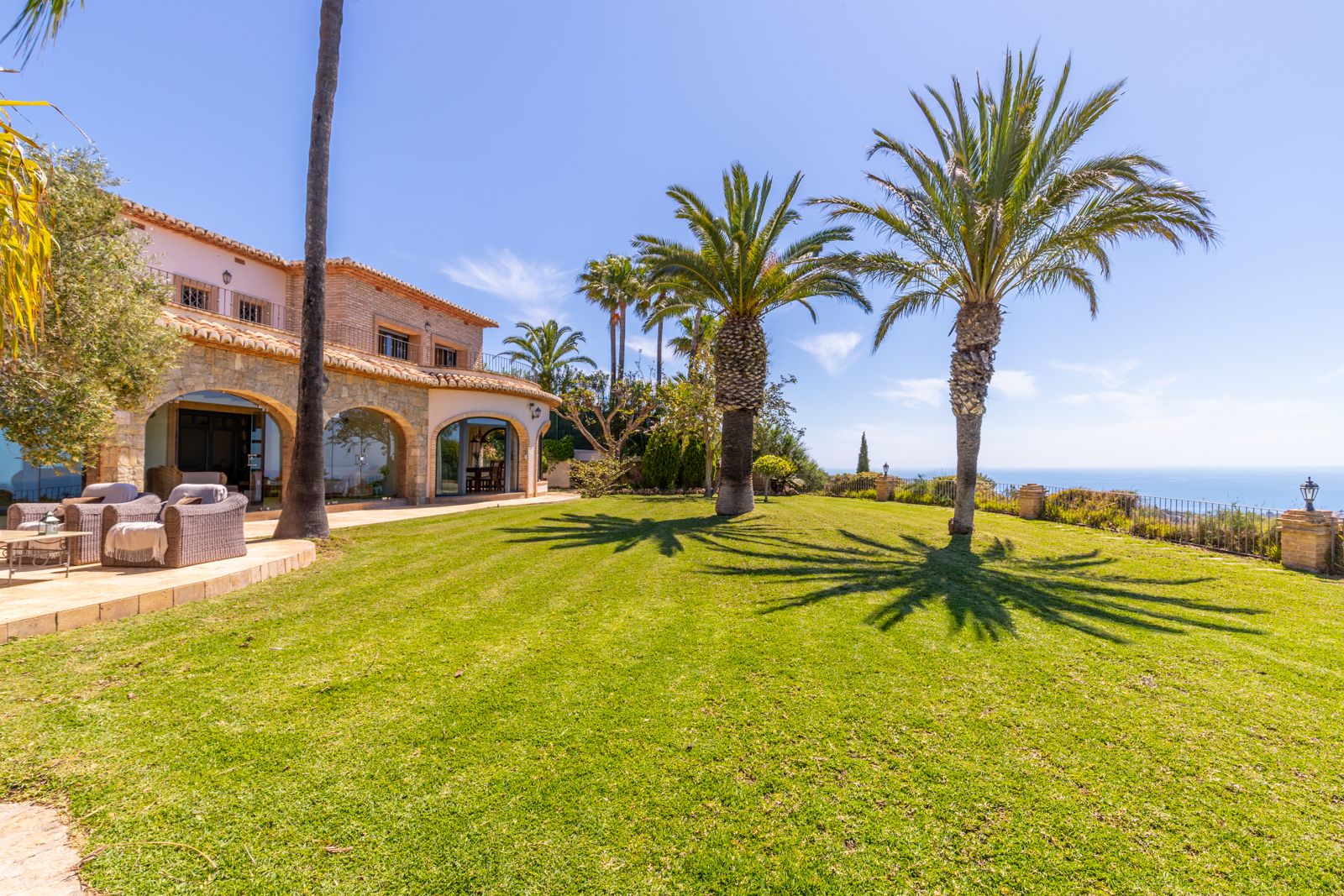 Vista del jardín con césped, palmeras y fachada de piedra con arcos de la finca mediterránea.