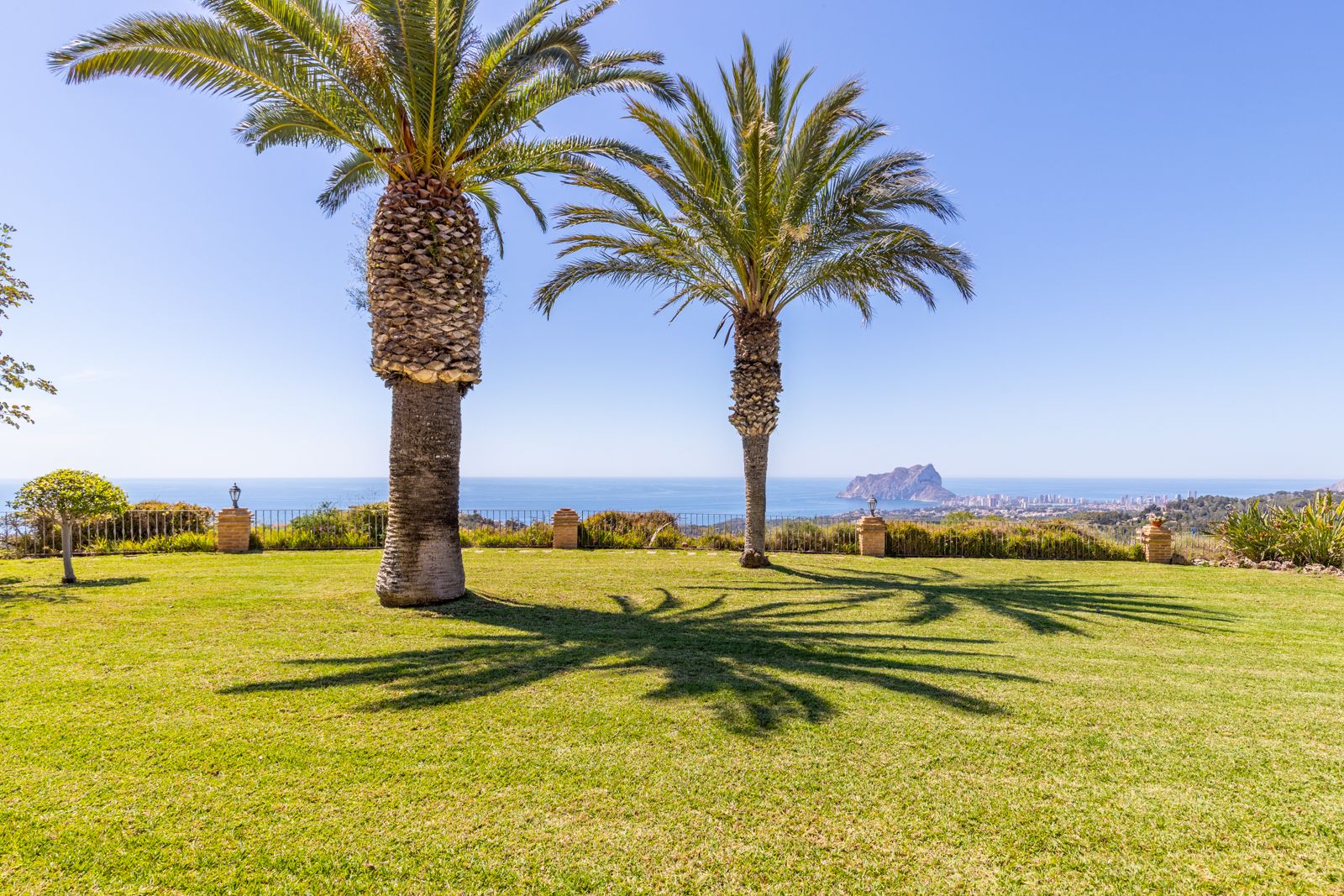 Vista del jardín con césped verde, palmeras y vistas al mar Mediterráneo y al Peñón de Ifach.