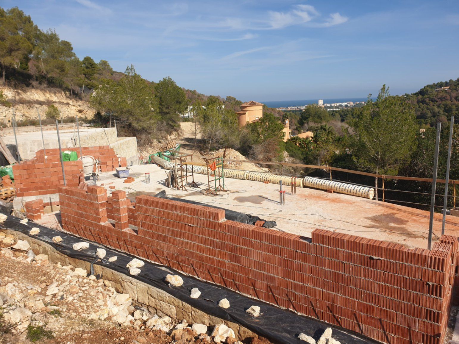 Estado actual de la construcción mostrando el levantamiento de muros de ladrillo cerámico sobre la cimentación, con vistas panorámicas al mar y al entorno natural de Jávea.