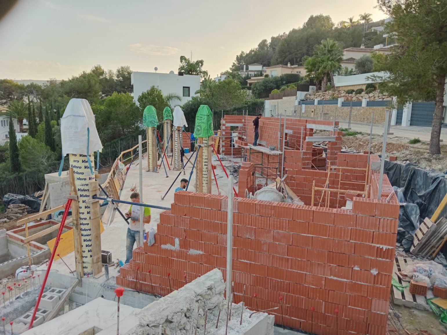 Estado actual de la obra mostrando el levantamiento de muros de ladrillo cerámico y pilares estructurales en una parcela elevada rodeada de vegetación mediterránea.