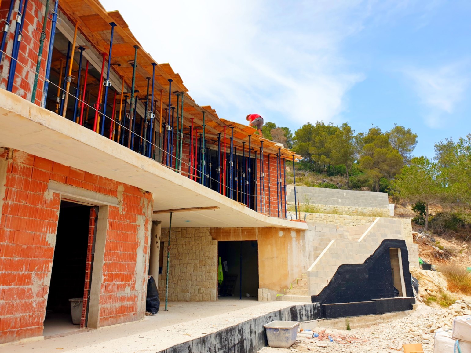 Vista exterior de la villa en fase de construcción, mostrando la estructura de ladrillo, el forjado superior con puntales y muros de piedra natural en la base.