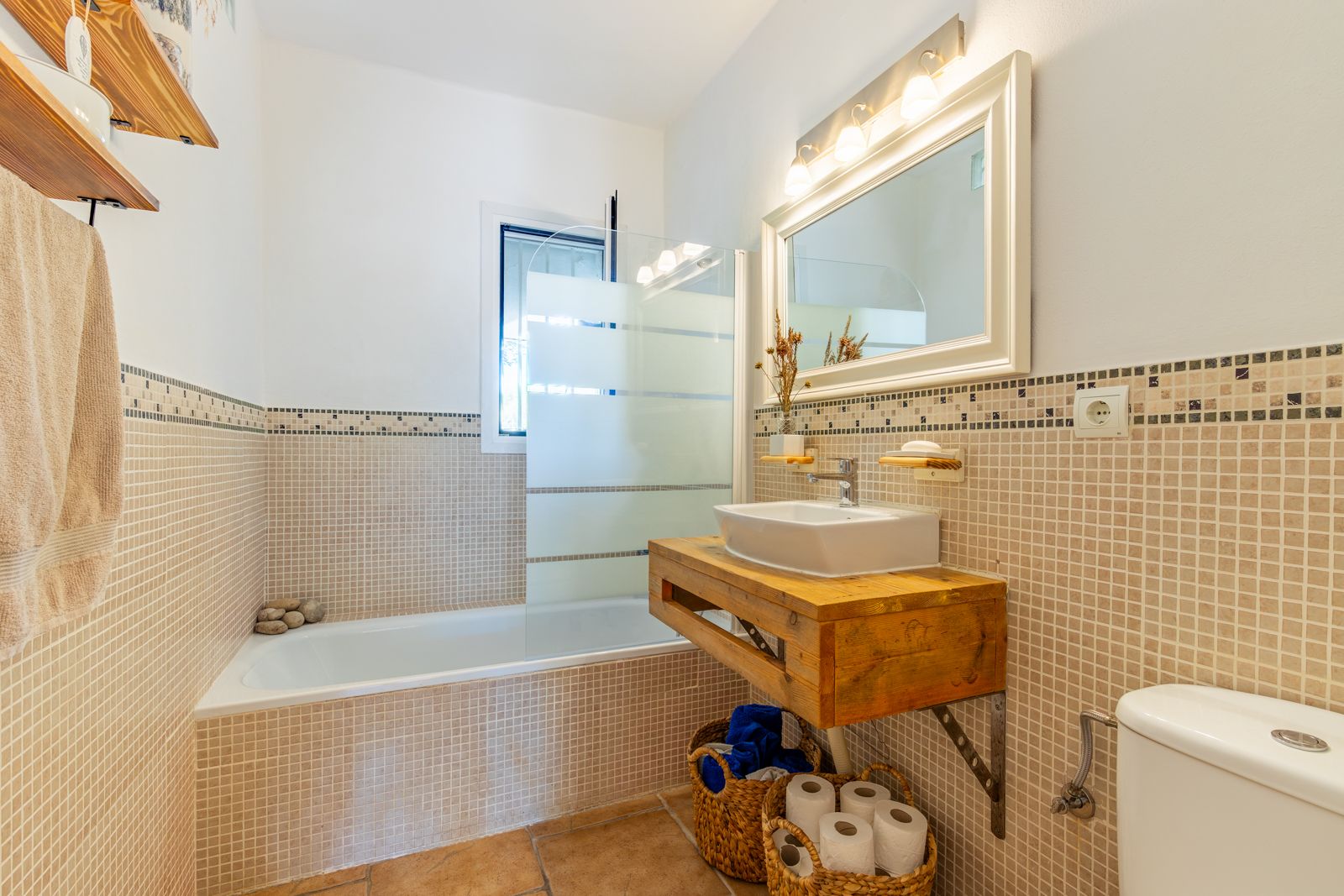 Baño de estilo rústico con encimera de madera maciza, lavabo sobre encimera, bañera con mampara de cristal y paredes revestidas en gresite beige.