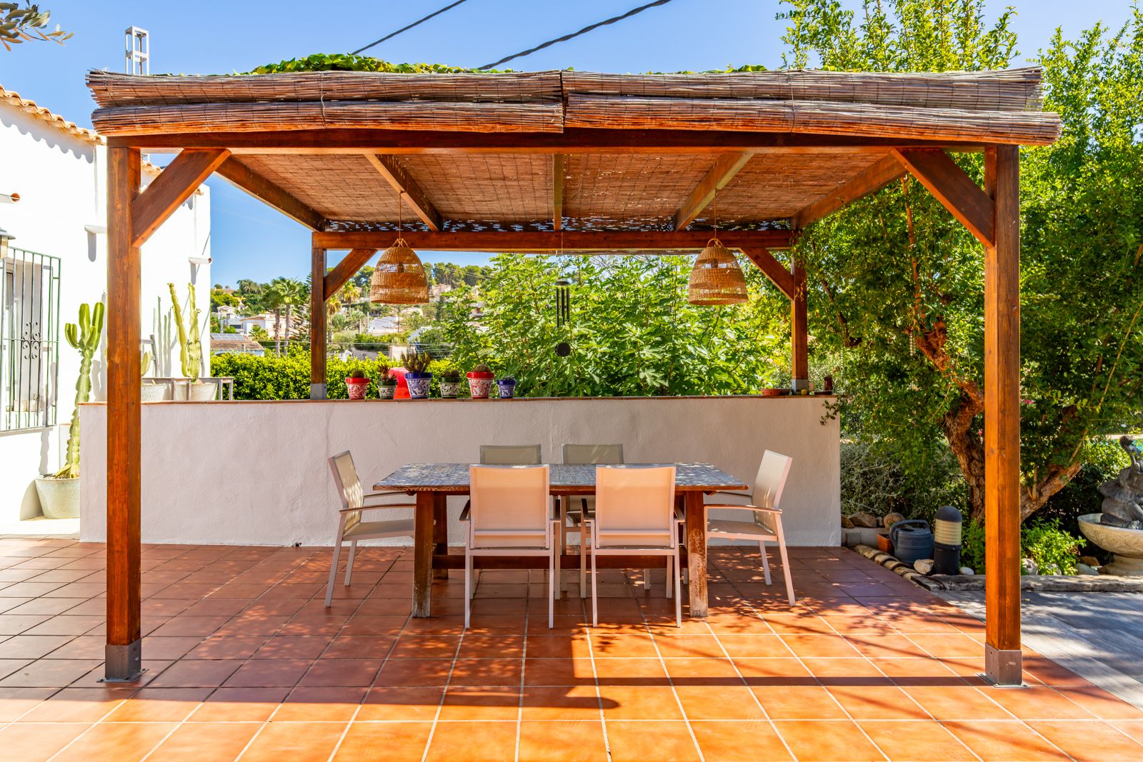 Comedor exterior bajo pérgola de madera con cubierta de cañizo, lámparas de mimbre y mesa de azulejos sobre suelo de terracota.