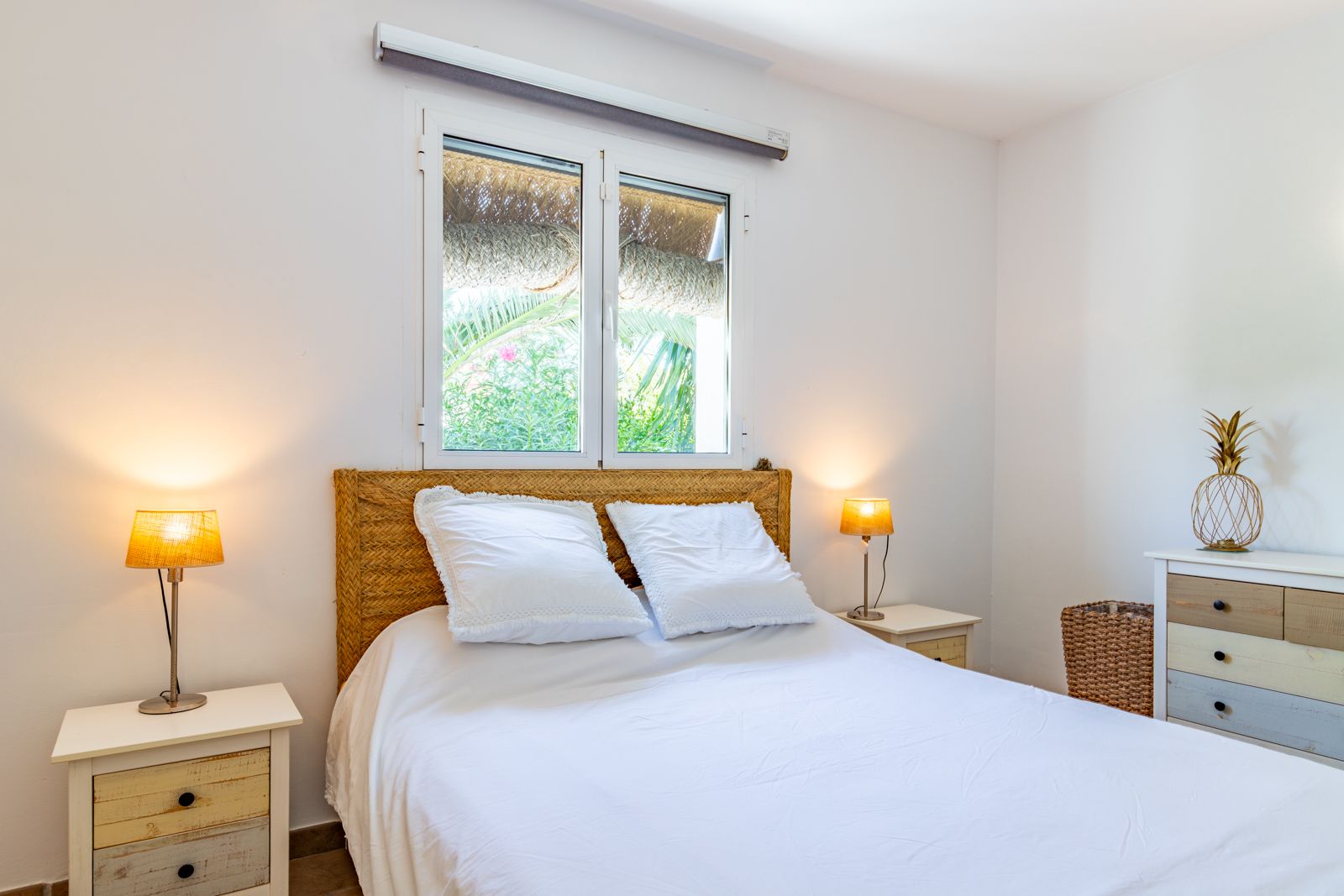 Dormitorio doble con cabecero de fibras naturales, mesitas de madera rústica y ventana con vistas al jardín. Un espacio luminoso de estilo mediterráneo.