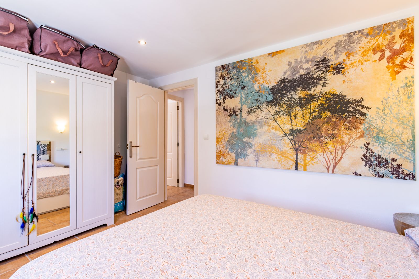 Dormitorio luminoso con armario blanco de tres puertas, espejo central y un gran mural decorativo de temática natural. Suelos cerámicos y puertas blancas.