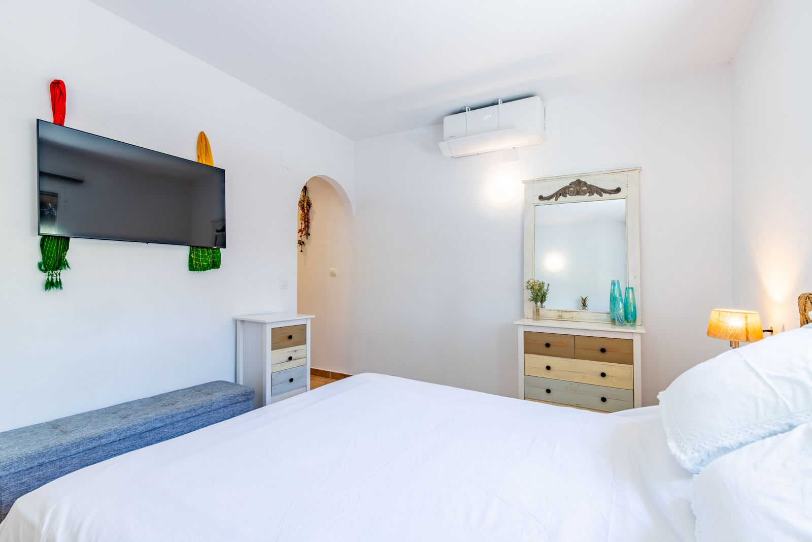 Dormitorio luminoso con cama doble, cómodas de estilo rústico, aire acondicionado y televisor de pantalla plana montado en la pared blanca.