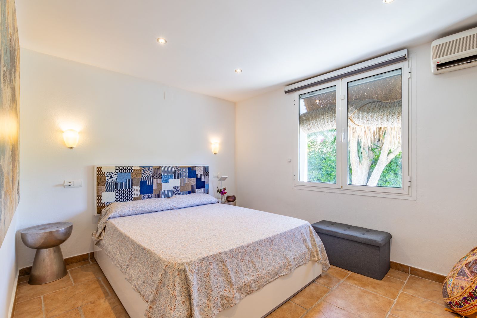 Dormitorio luminoso con suelos de terracota, cabecero de patchwork, aire acondicionado y ventana con vistas a la vegetación exterior.