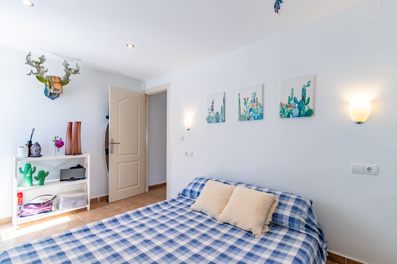 Dormitorio luminoso con suelos de terracota, cama con textiles a cuadros y decoración temática. Incluye iluminación empotrada y apliques de pared cálidos.
