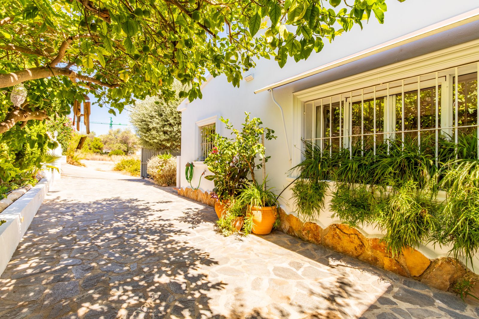 Fachada blanca con zócalo de piedra y ventanas enrejadas, rodeada de vegetación mediterránea y un camino de piedra natural bajo la sombra de los árboles.