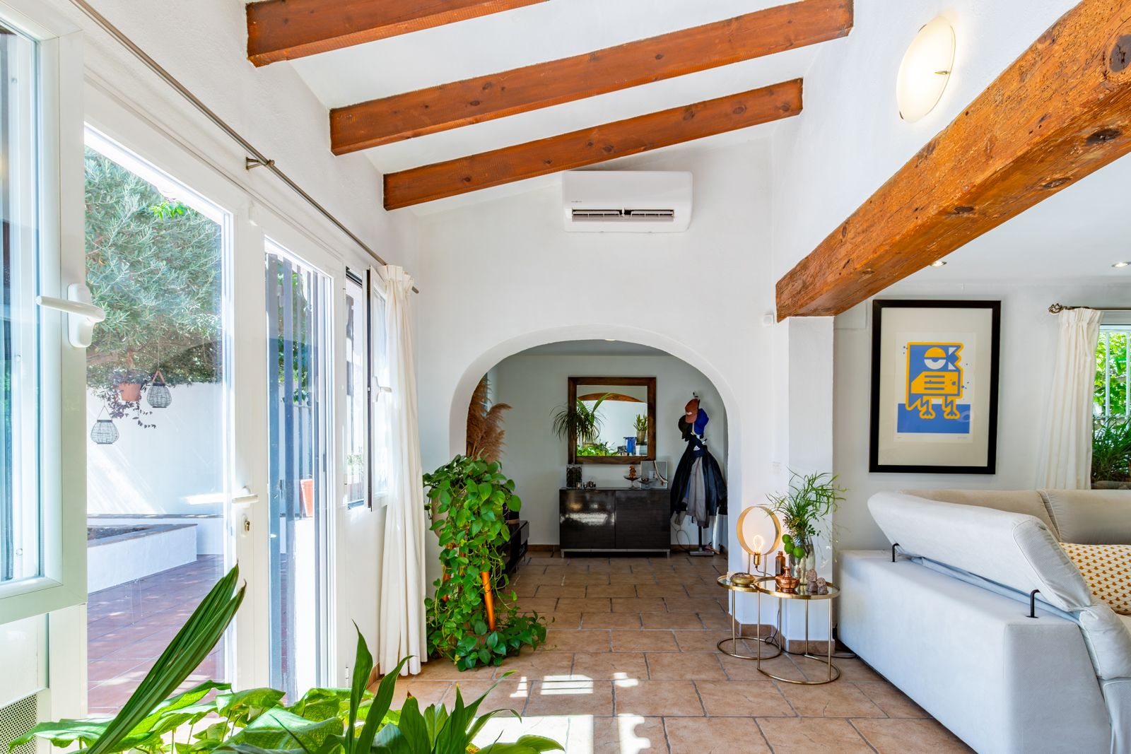 Salón luminoso con vigas de madera vistas, suelos de terracota y acceso directo a la terraza. Un arco comunica con el recibidor bajo un techo inclinado con aire acondicionado.