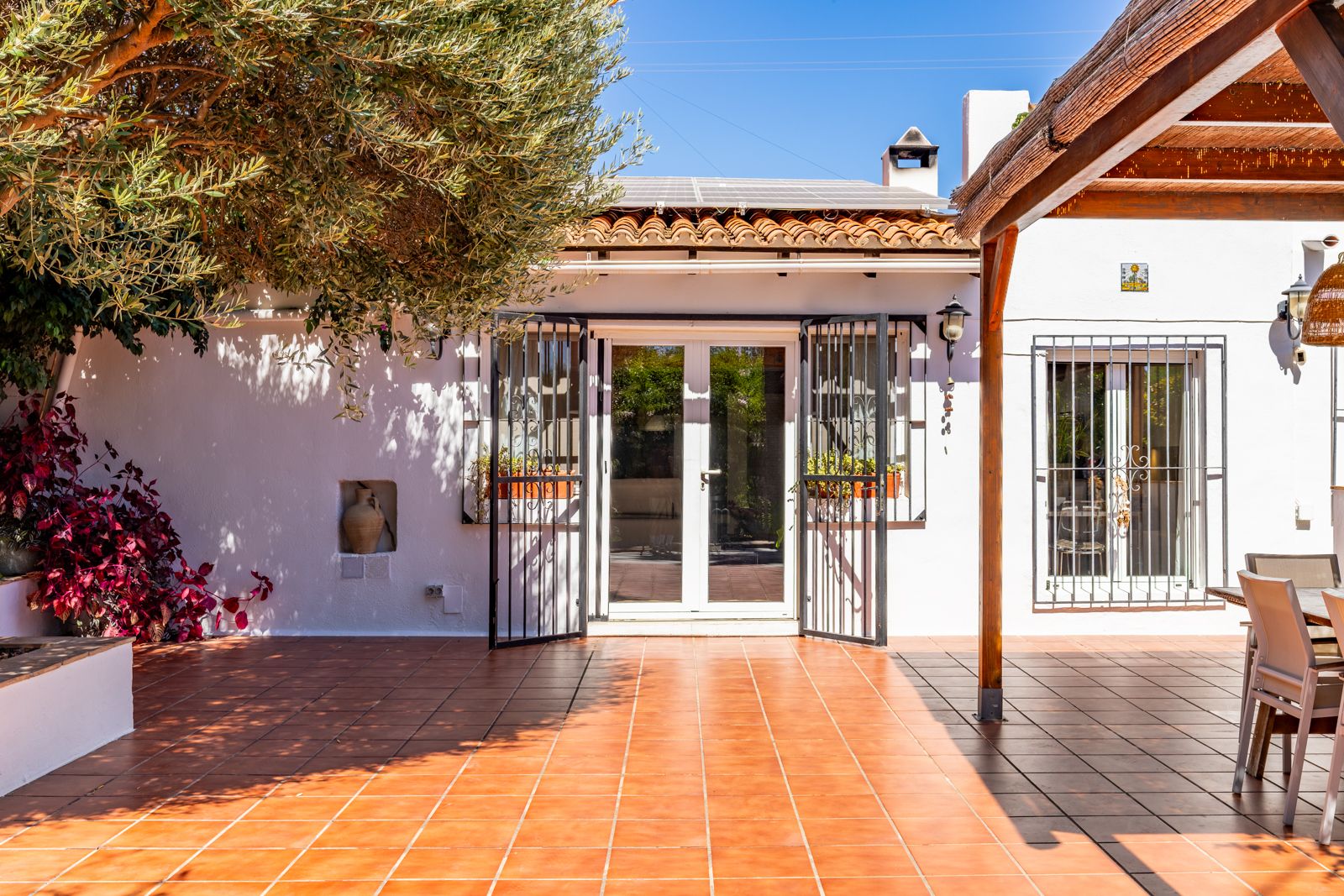Terraza exterior con suelo de baldosas de barro, pérgola de madera y paneles solares. La fachada blanca destaca con rejas de hierro negro y un olivo que proporciona sombra natural.