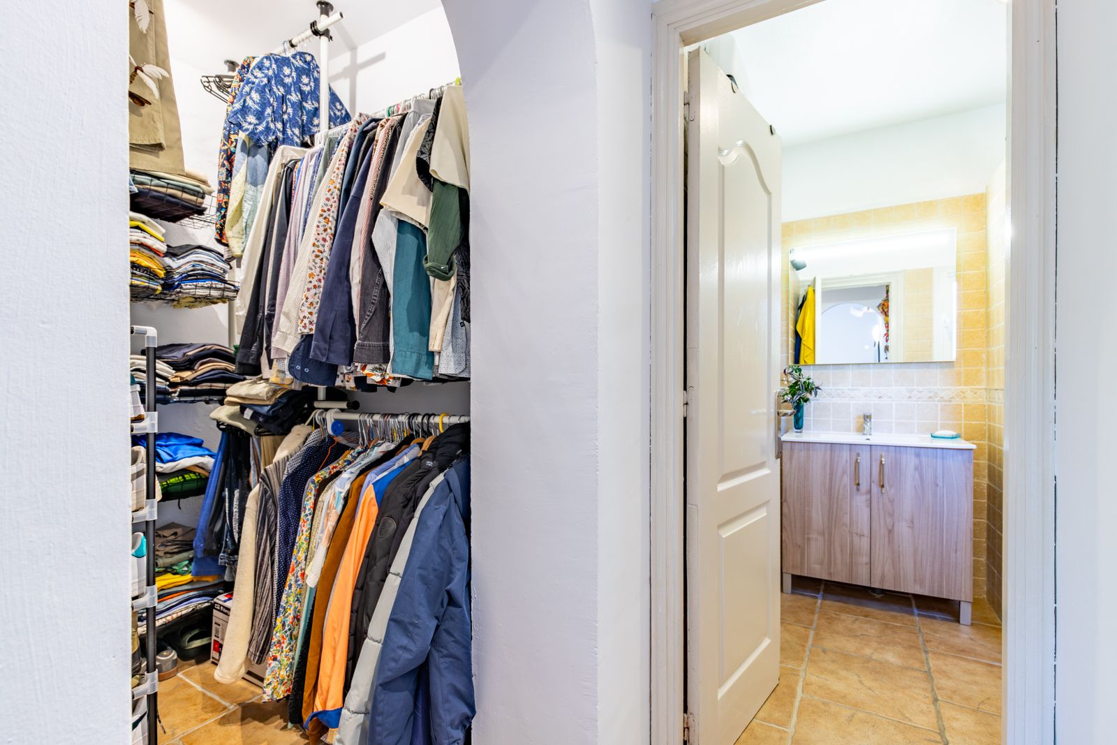 Vestidor abierto con estanterías y barras de colgar, comunicado con un baño que incluye mueble de madera, espejo retroiluminado y suelos cerámicos.