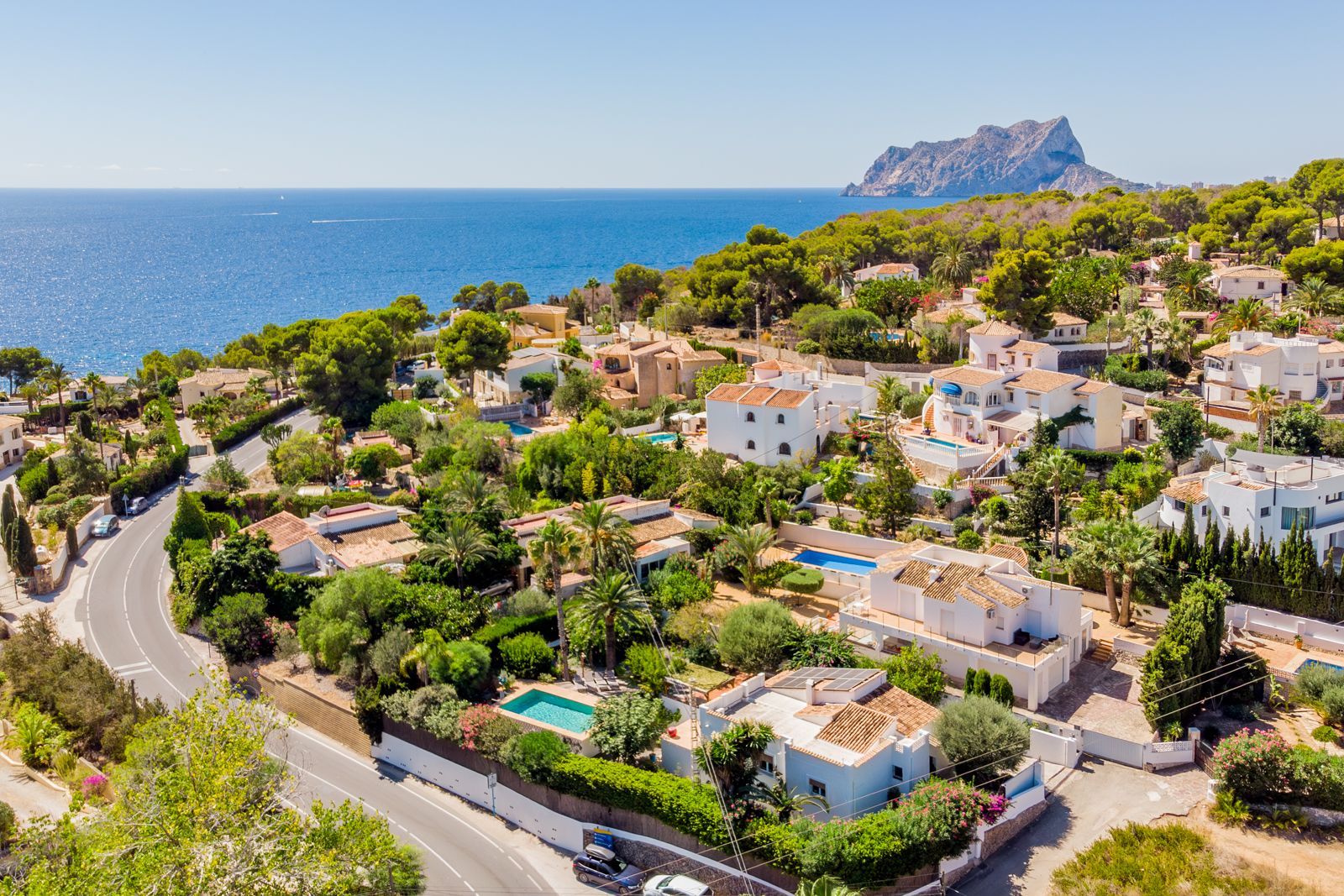 Vista aérea de la costa de Baladrar con el Peñón de Ifach al fondo. La zona residencial destaca por su proximidad al mar y su entorno rodeado de vegetación mediterránea.
