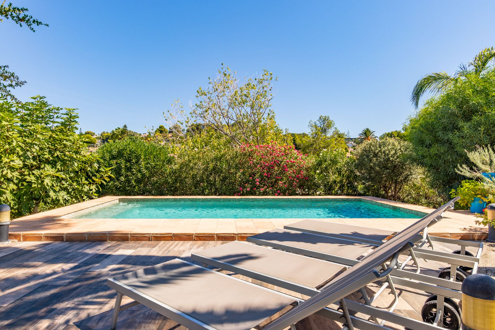 Zona de piscina privada con solárium de gres cerámico efecto madera, rodeada de vegetación mediterránea madura y equipada con hamacas modernas.