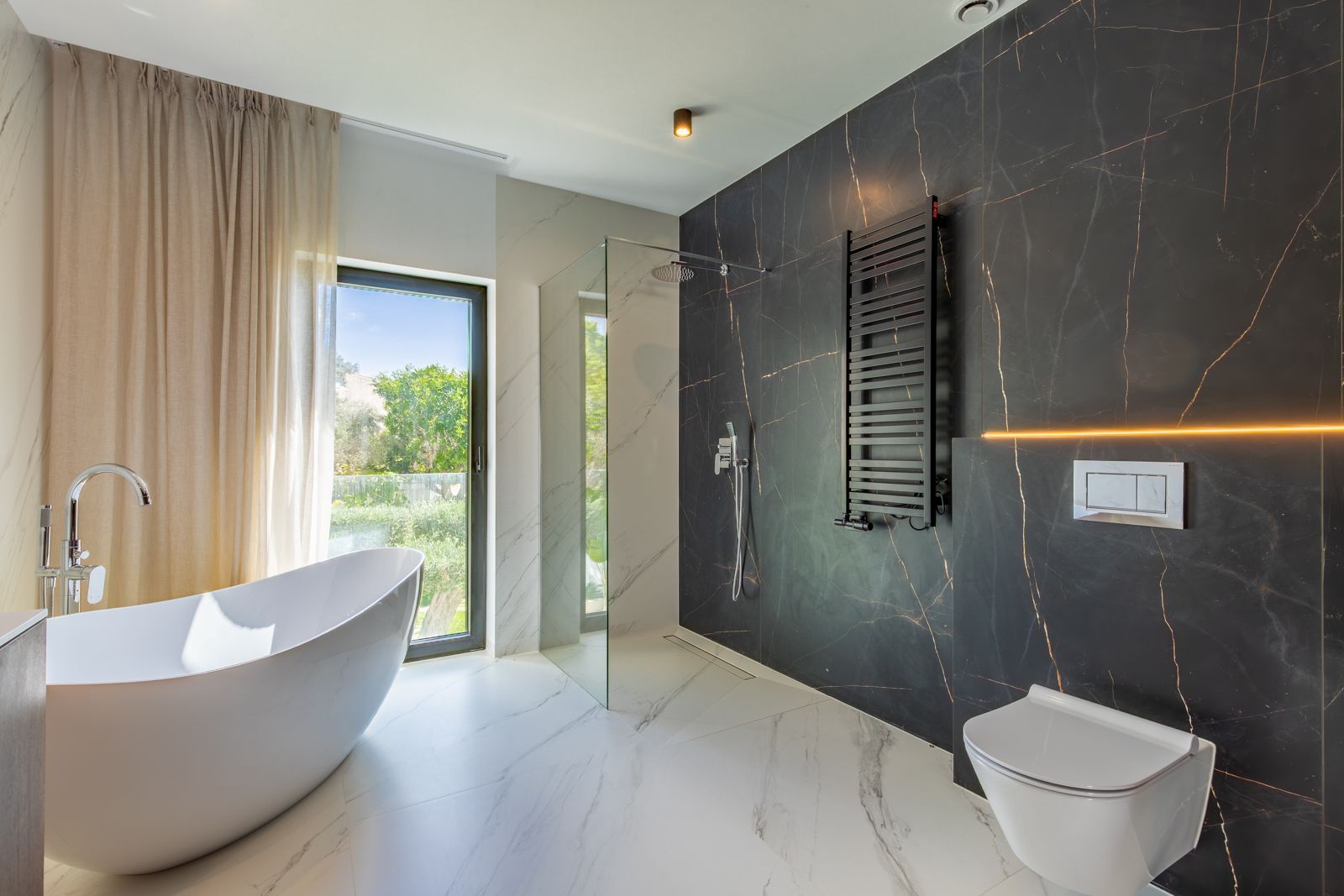 Baño de diseño con bañera exenta, ducha italiana y revestimientos de gres porcelánico en contraste. Incluye inodoro suspendido, radiador toallero negro y vistas al jardín.