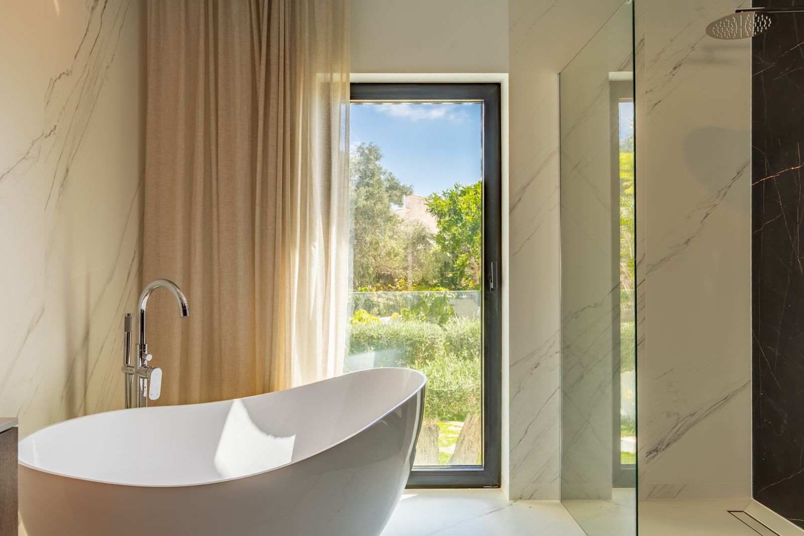 Baño de diseño con bañera exenta, grifería de pie y ducha italiana. Revestimientos de gres porcelánico efecto mármol y ventanal con vistas al jardín mediterráneo.