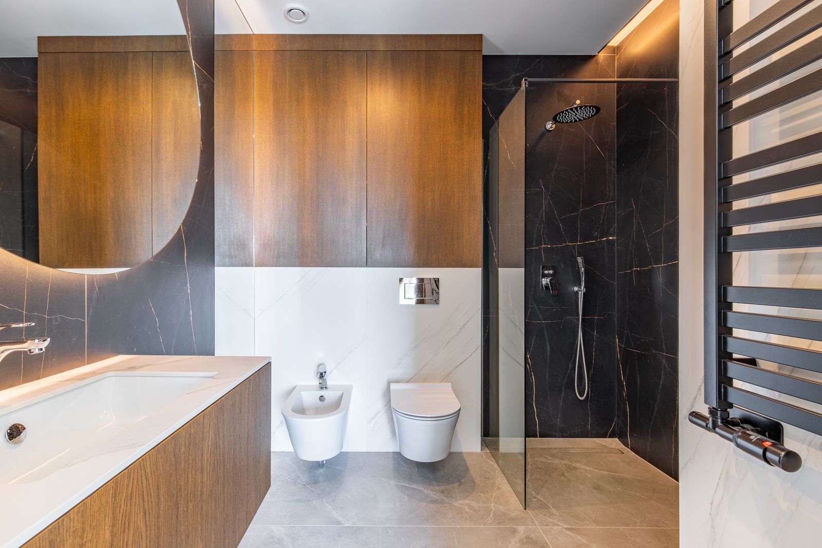 Baño moderno con revestimiento de porcelánico oscuro, ducha italiana, sanitarios suspendidos y muebles de roble natural bajo un gran espejo circular retroiluminado.
