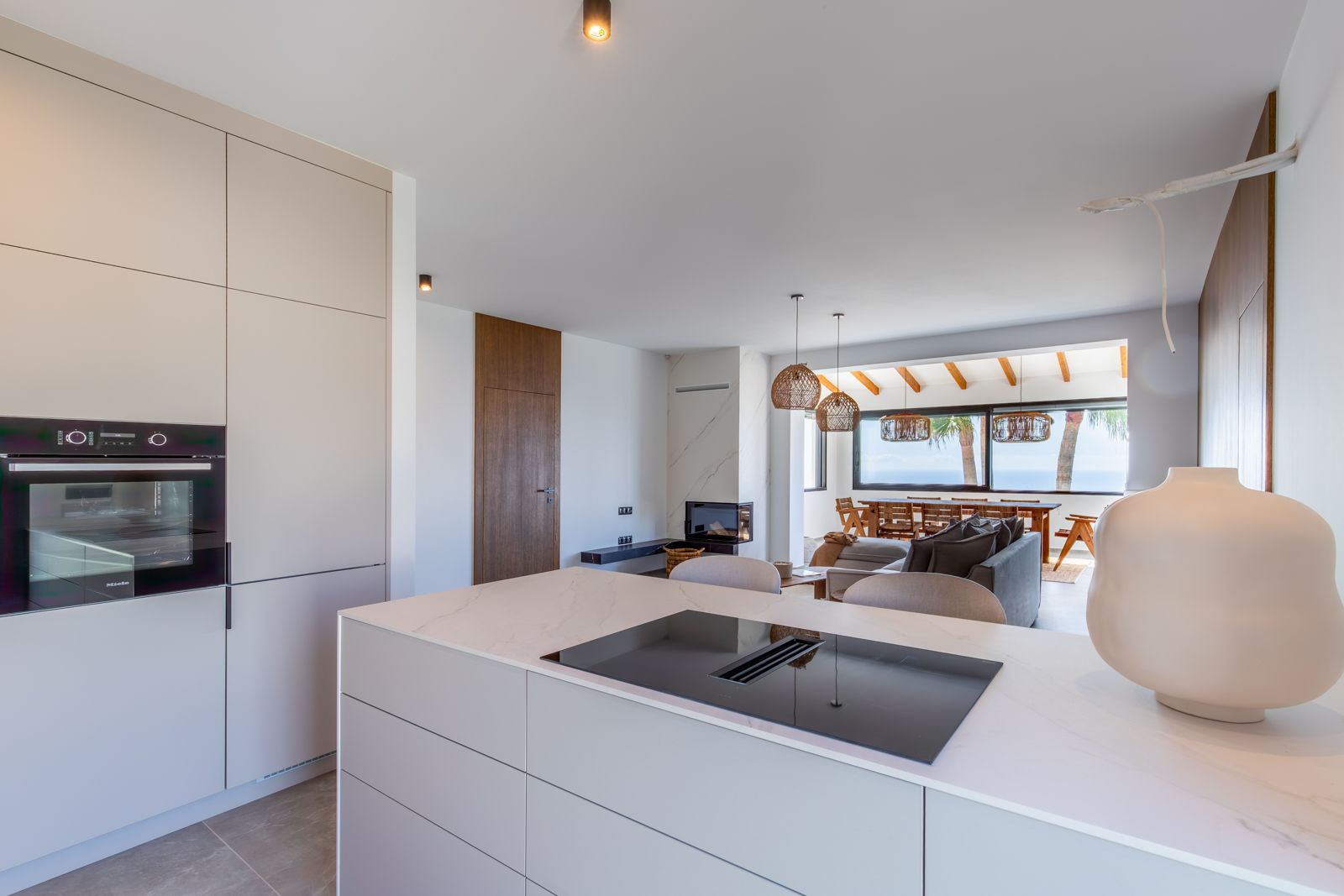 Cocina de diseño abierta al salón con isla de piedra sinterizada, placa de inducción integrada y electrodomésticos Miele, ofreciendo vistas al mar a través del comedor.