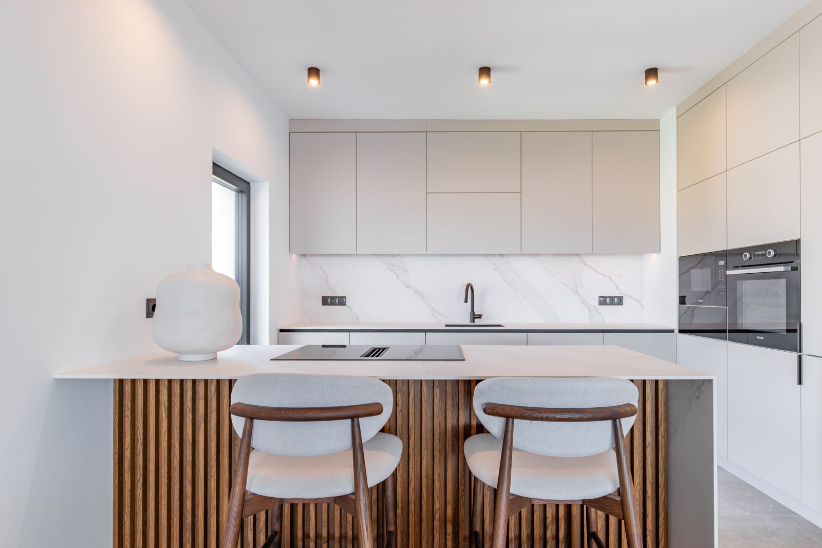 Cocina de diseño con isla central revestida en listones de roble, encimera de piedra sinterizada y electrodomésticos Miele integrados en mobiliario minimalista.