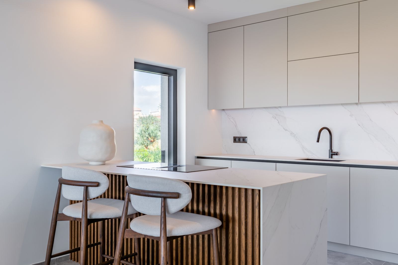 Cocina de diseño con isla de piedra sinterizada efecto mármol, panelado de listones de madera y taburetes ergonómicos. Incluye placa de inducción integrada y grifería minimalista.