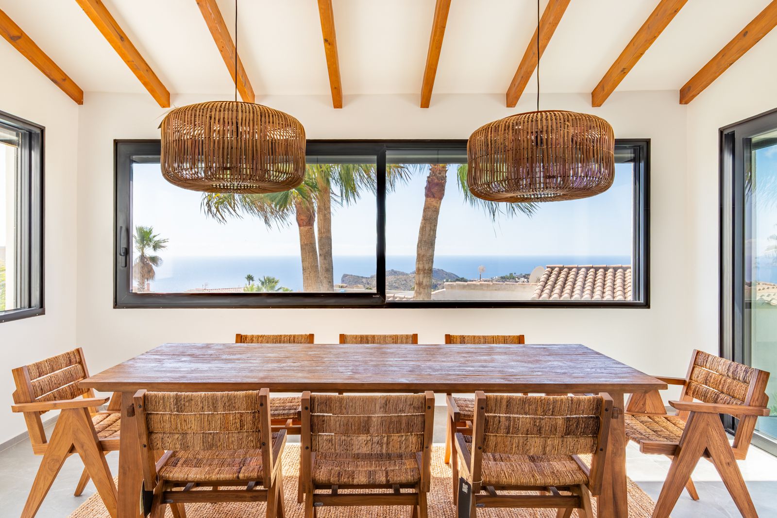 Comedor con vigas de madera, mesa maciza y lámparas de mimbre. El gran ventanal enmarca vistas directas al mar Mediterráneo y las palmeras del entorno.