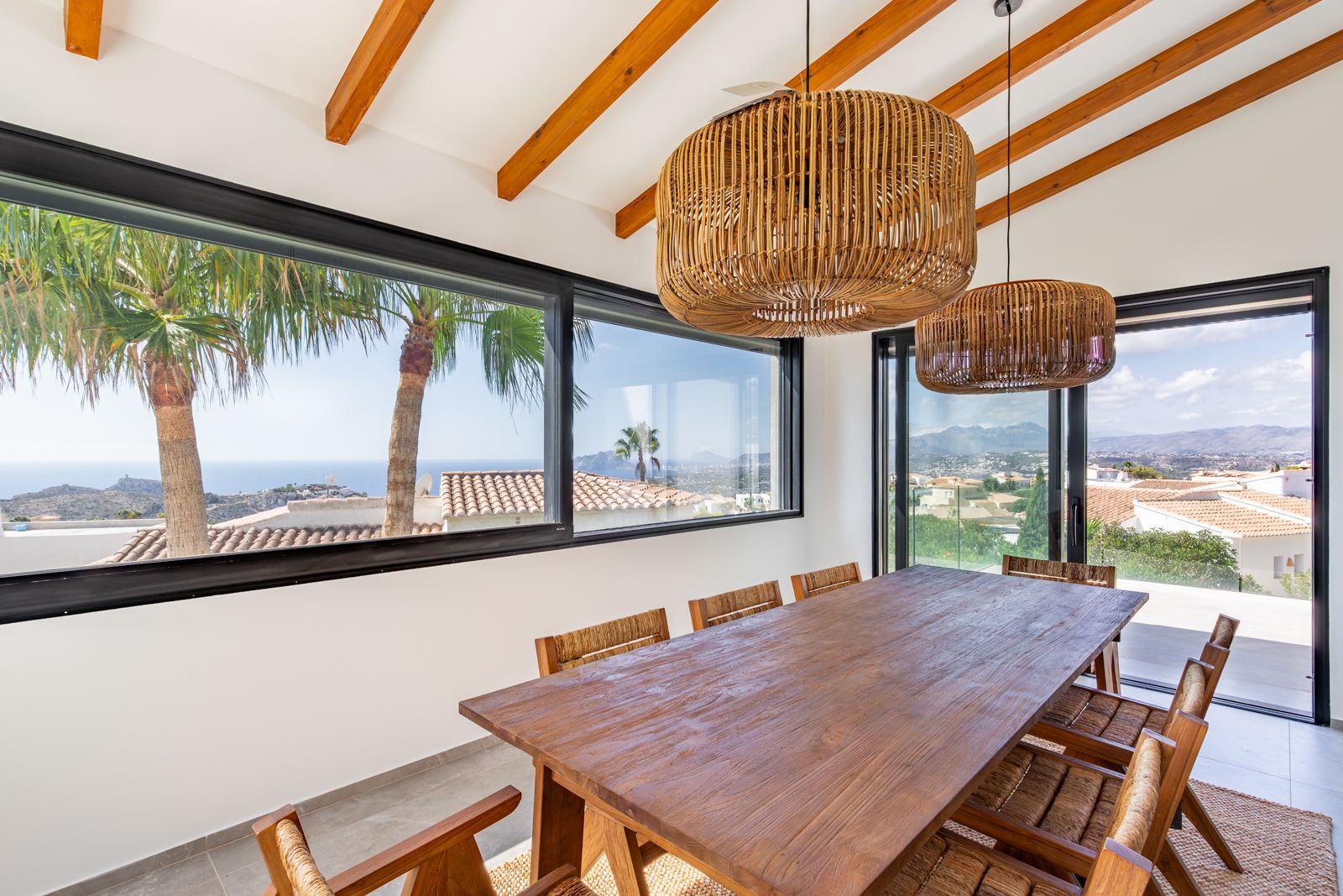 Comedor luminoso con vigas de madera, mesa de madera maciza y lámparas de fibra natural. Grandes ventanales ofrecen vistas panorámicas al mar Mediterráneo y las montañas.