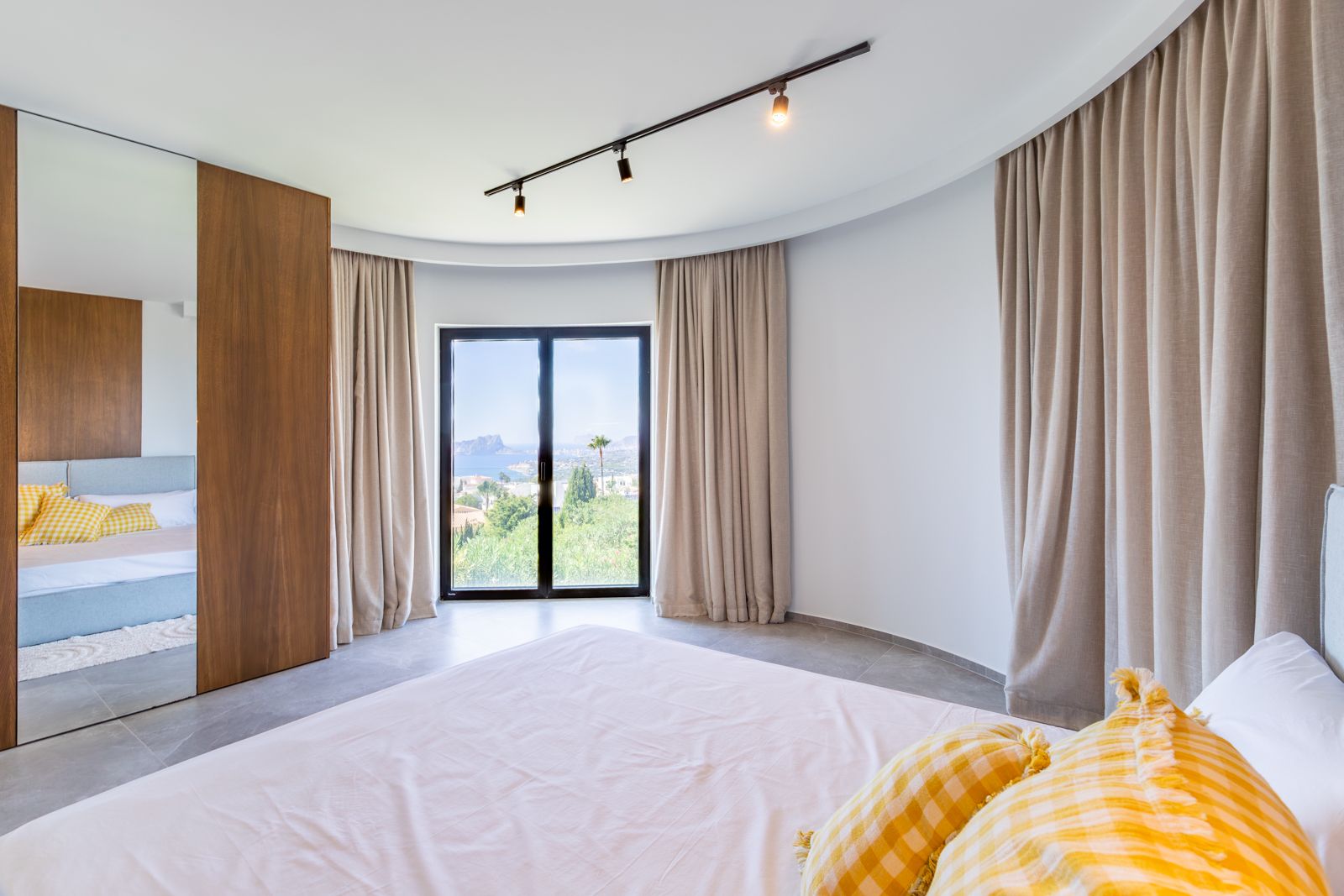 Dormitorio con pared curva, armario empotrado en roble natural y vistas al mar a través de ventanales de triple acristalamiento con cortinas de suelo a techo.