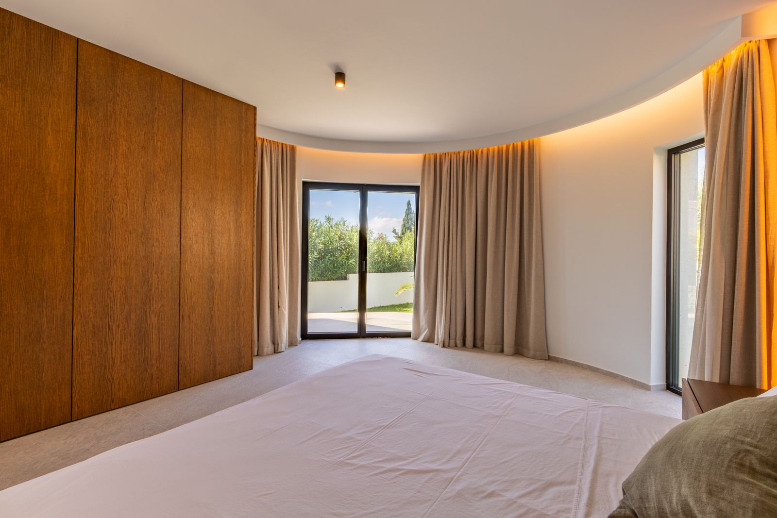 Dormitorio con pared curva, armarios de roble natural e iluminación LED indirecta. Grandes ventanales conectan el espacio con el jardín exterior.