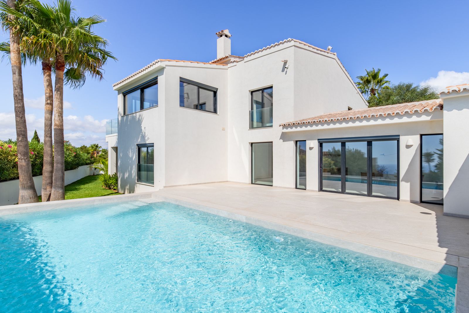 Exterior de la villa con piscina de cloración salina y amplia terraza de gres porcelánico. La fachada blanca destaca por su carpintería de aluminio y ventanales de triple acristalamiento.