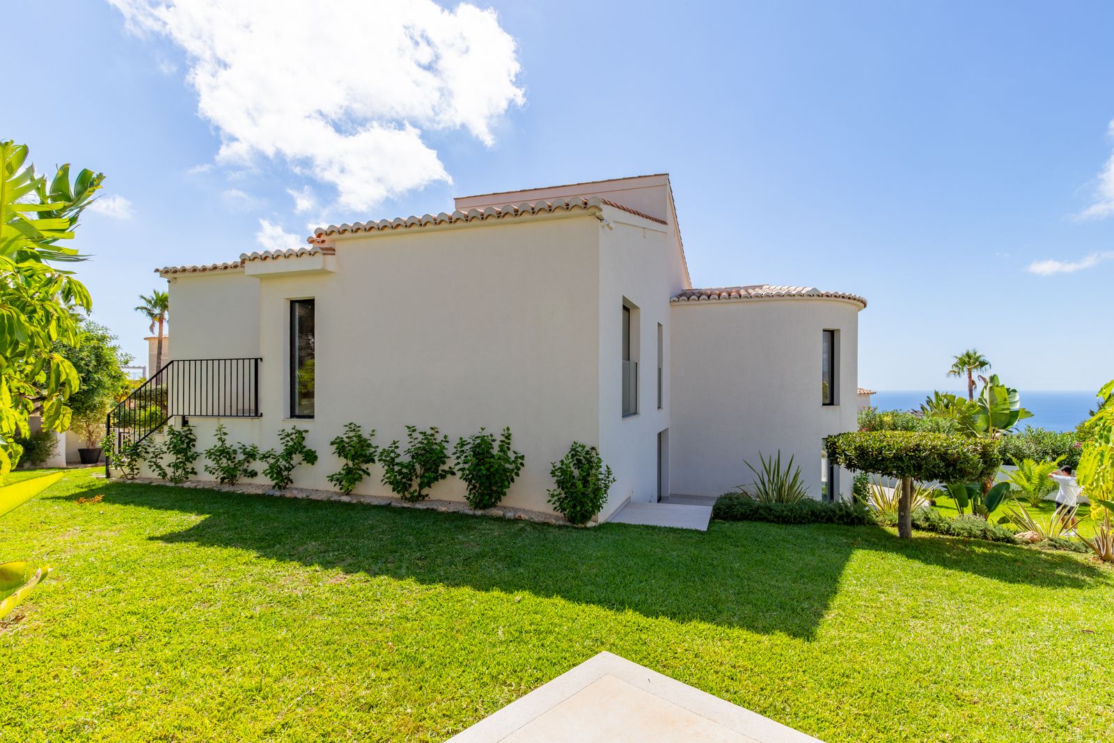 Fachada blanca de diseño mediterráneo con tejado de teja, jardín de césped natural y vistas al mar desde una zona elevada con vegetación tropical.