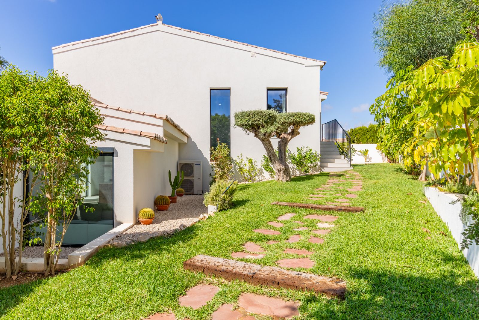 Jardín mediterráneo con césped cuidado, olivo ornamental y sendero de piedra natural que conduce a la fachada blanca de la villa con ventanales verticales.