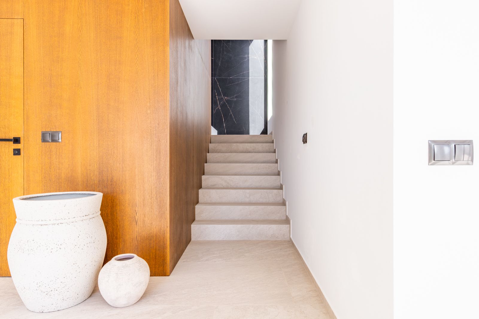 Pasillo con escalera de gres porcelánico, pared revestida en chapa de roble natural y fondo de piedra sinterizada negra con veteado blanco.