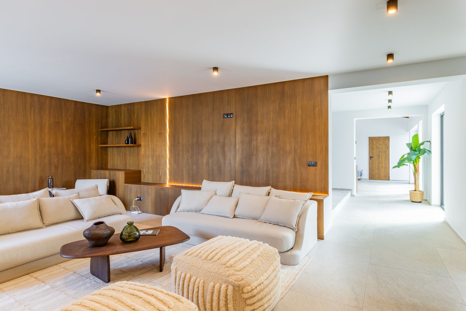 Salón con paneles de roble natural e iluminación LED integrada. Mobiliario en tonos neutros sobre suelos de gres porcelánico y techos con focos técnicos cilíndricos.