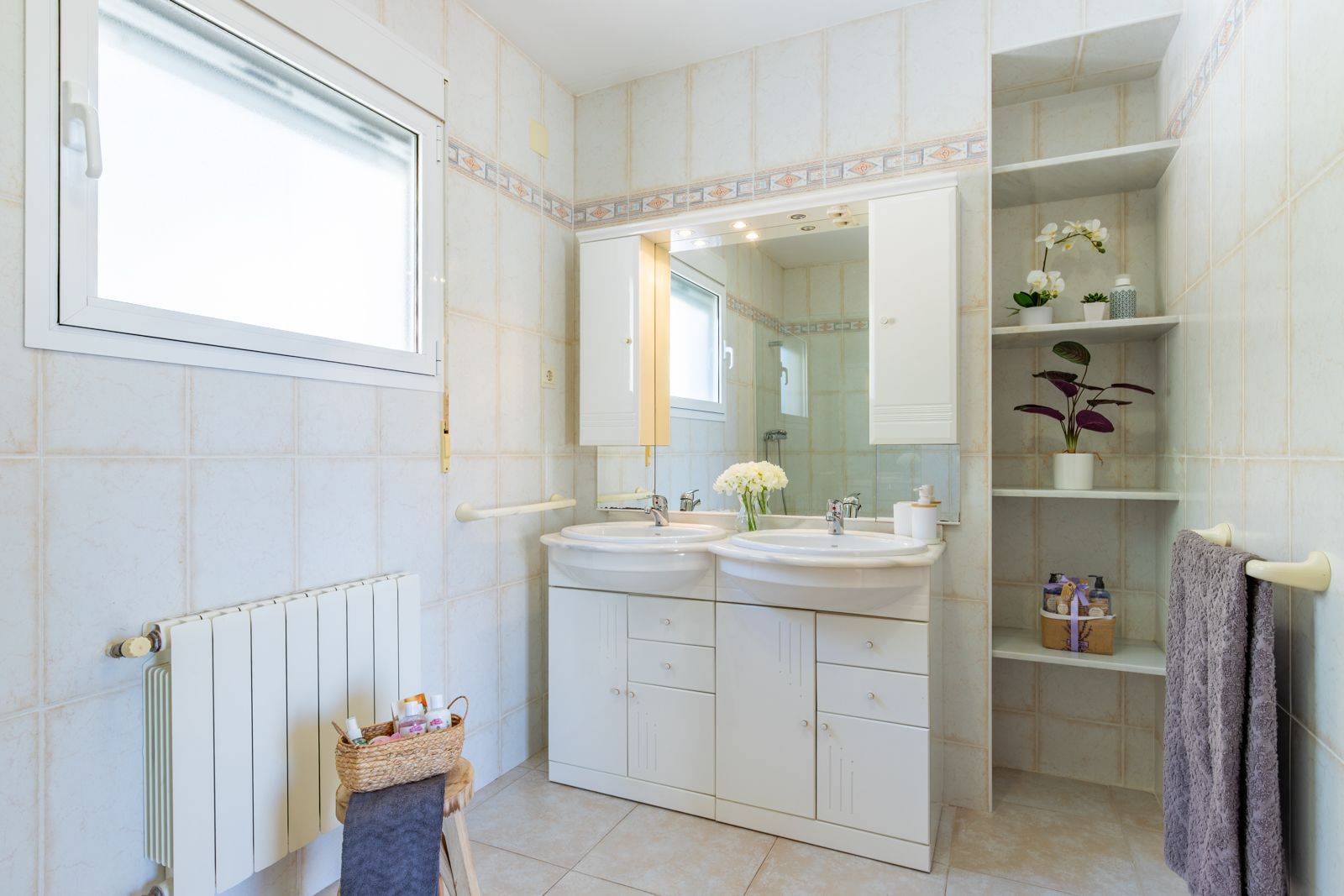 Baño con lavabo de doble seno, mueble blanco con amplio almacenaje, espejo iluminado y estantería de obra. Paredes alicatadas con cenefa decorativa y radiador integrado.