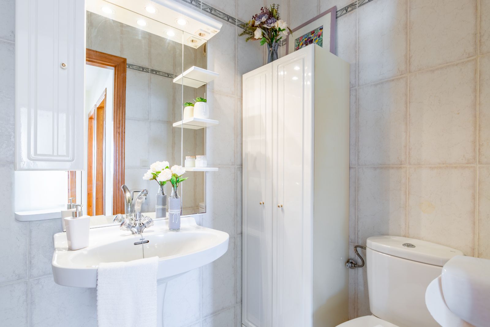 Baño funcional con lavabo de pedestal, espejo con iluminación integrada y estanterías de cristal, complementado por un armario de almacenamiento blanco y azulejos en tonos neutros.