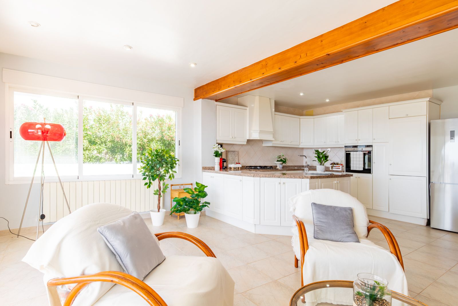Cocina abierta con armarios blancos y encimera de granito, integrada en un salón luminoso con vigas de madera vista y amplios ventanales al jardín.