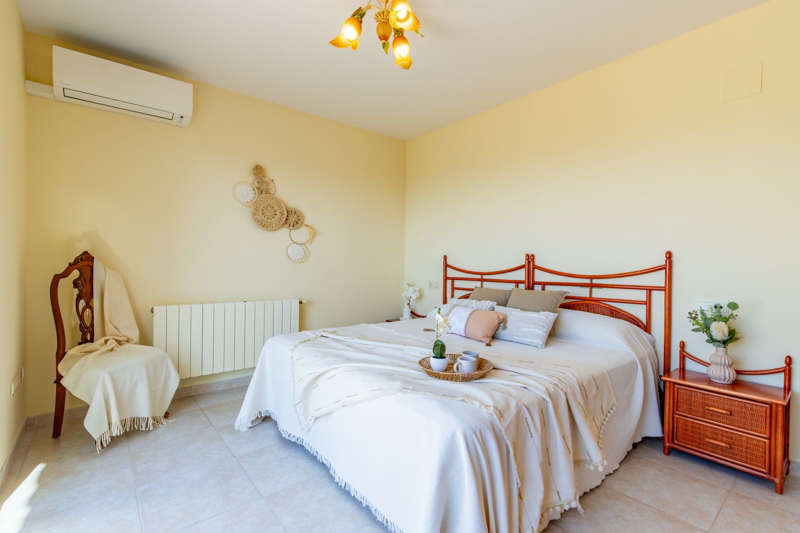 Dormitorio luminoso con cama doble, cabecero de madera tallada, mesita de noche a juego, aire acondicionado split, radiador y suelos de gres en tonos neutros.