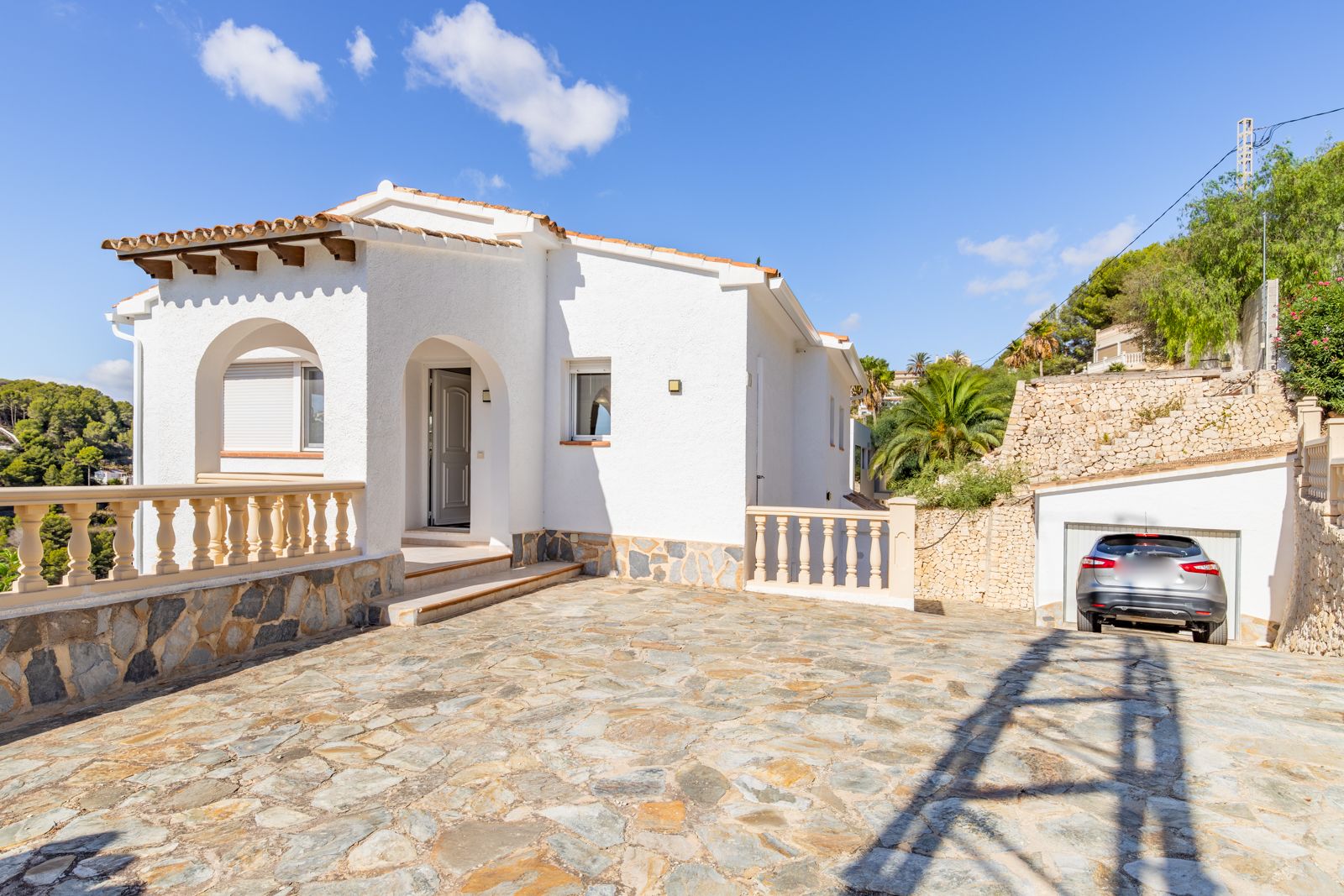 Fachada mediterránea con entrada porticada, vigas de madera y amplio patio de piedra natural que conduce al garaje privado y acceso principal.