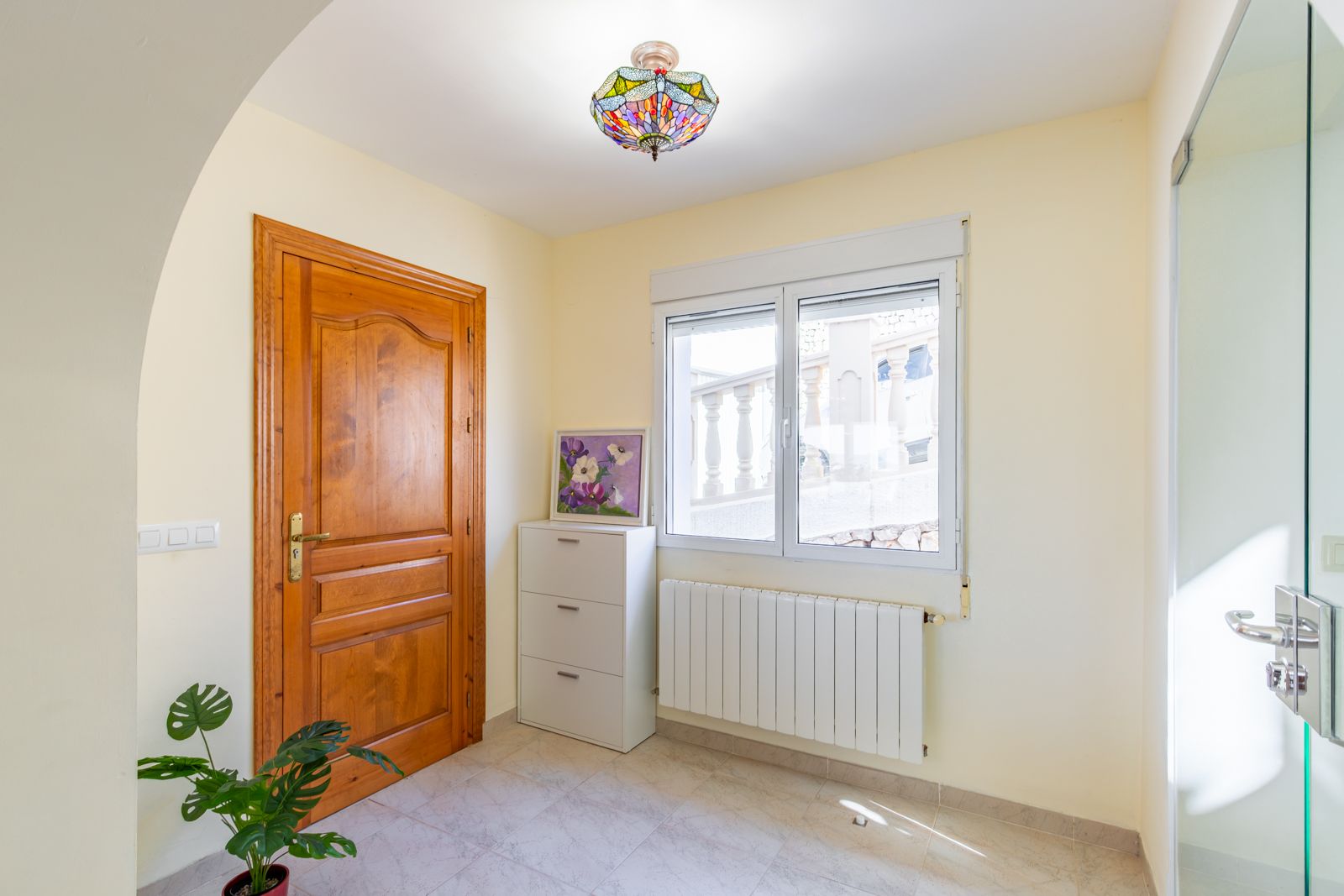 Hall de entrada con puerta de madera maciza, radiador de calefacción central y una distintiva lámpara de techo estilo Tiffany sobre suelos cerámicos claros.