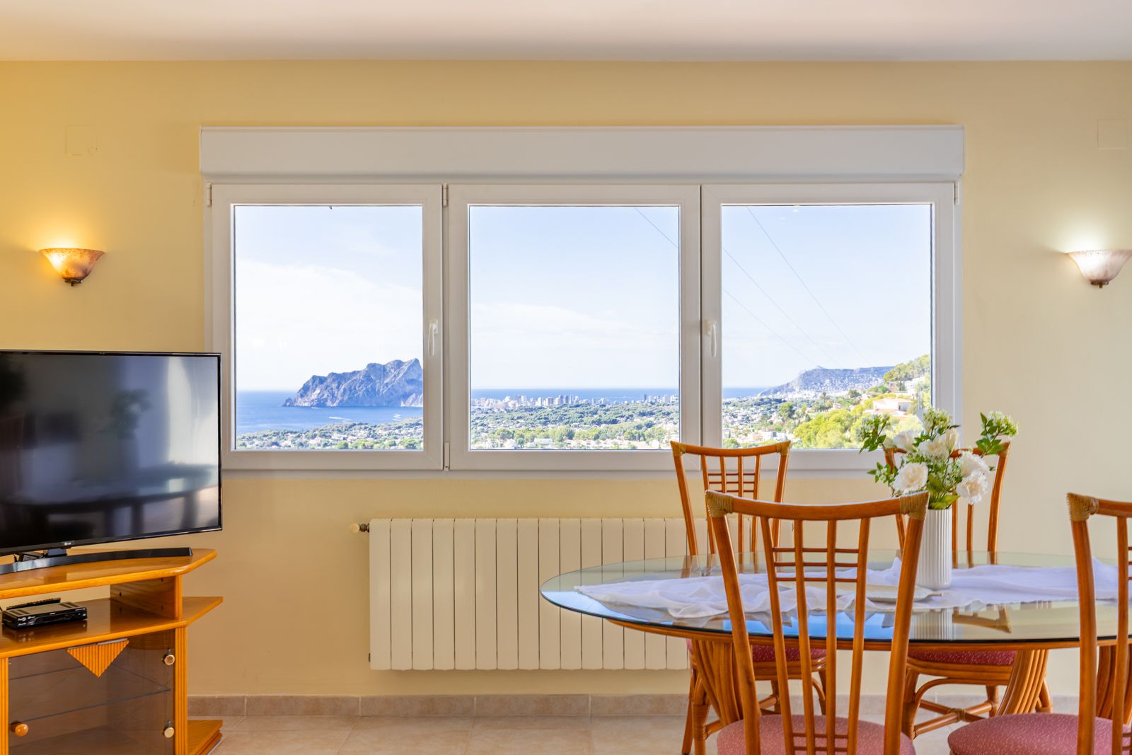 Salón comedor con amplios ventanales que ofrecen vistas panorámicas al mar y al Peñón de Ifach. Incluye calefacción central, mesa de cristal y mobiliario de madera.