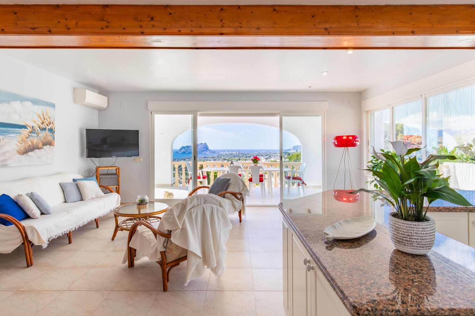 Salón luminoso con vigas de madera vistas, cocina integrada con encimera de granito y acceso directo a una terraza con vistas panorámicas al mar y al Peñón de Ifach.